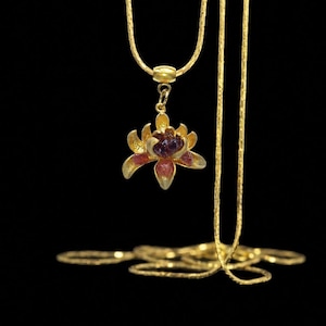 May include: A gold-toned necklace featuring a pendant shaped like a blooming flower. The flower petals are crafted from gold-colored metal, with a cluster of small, deep purple and reddish-pink raw gemstones set in the center of the blossom.