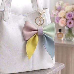 Könnte beinhalten: Eine weiße Handtasche mit einem mehrfarbigen Schleifen-Schlüsselanhänger am Griff. Die Schleife besteht aus vier verschiedenen Segmenten in Pastellrosa, Hellgrün, Gelb und Blau, die durch ein zentrales lavendelfarbenes Band und einen Goldring verbunden sind.