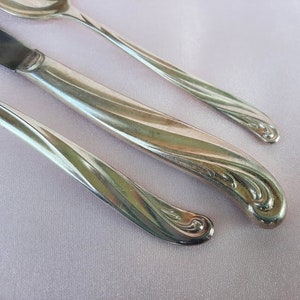 May include: Close-up of antique silverware set on a light pink surface. The set includes a knife, spoon, and fork, all with a silver finish and ornate, swirling handle designs. The silverware is arranged diagonally, showcasing the elegant details.