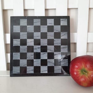 May include: A square black and gray marble chess board with a checkerboard pattern. The board is next to a red apple. The chess board is made of marble and has a polished finish.