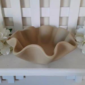 May include: A beige, ruffled-edge bowl. The bowl has a wavy, scalloped rim and a smooth interior. The bowl is sitting on a white shelf, with white flowers and a white lattice background.