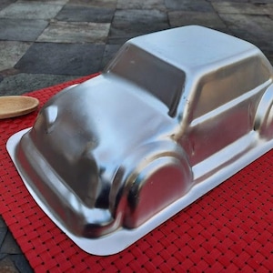 May include: A silver-colored car-shaped cake pan. The pan is made of metal and has a rectangular base. A wooden spoon and a metal spoon are on the red placemat.
