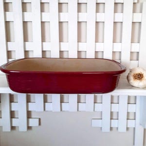 May include: A rectangular, burgundy-colored ceramic baking dish with rounded edges. The interior is a light tan color. A whole garlic bulb sits to the right of the dish on a white shelf against a white slatted background.