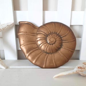 May include: A gold-colored metal seashell with a detailed spiral design. The seashell is on a white surface.