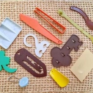 May include: A collection of various plastic kitchen tools and accessories in a variety of colors, including orange, green, brown, and yellow. The items are arranged on a woven, beige surface.