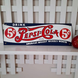 May include: Vintage Pepsi-Cola sign with red and white lettering on a blue background. The sign reads "DRINK 5&cent; Pepsi-Cola 5&cent; AMERICA'S BIGGEST NICKEL'S WORTH". A red apple sits to the right of the sign.