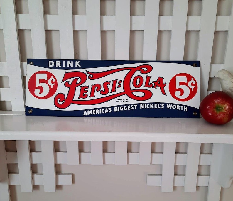 May include: Vintage Pepsi-Cola sign with red and white lettering on a blue background. The sign reads "DRINK 5&cent; Pepsi-Cola 5&cent; AMERICA'S BIGGEST NICKEL'S WORTH". A red apple sits to the right of the sign.