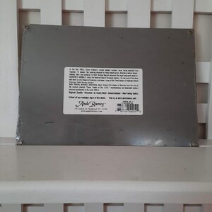 May include: A rectangular, gray metal sign with text and a white label. The label includes the text "Ande Rooney" and a website address. The sign is resting on a white slatted surface.