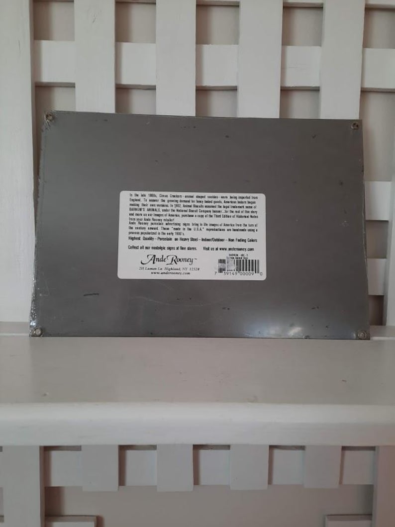 May include: A rectangular, gray metal sign with text and a white label. The label includes the text "Ande Rooney" and a website address. The sign is resting on a white slatted surface.