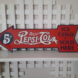 May include: Vintage Pepsi-Cola arrow-shaped sign in red with white and blue accents. The sign reads "Drink Pepsi-Cola, Delicious-Healthful" and "Ice Cold Sold Here." The sign also features the price "5&cent;".