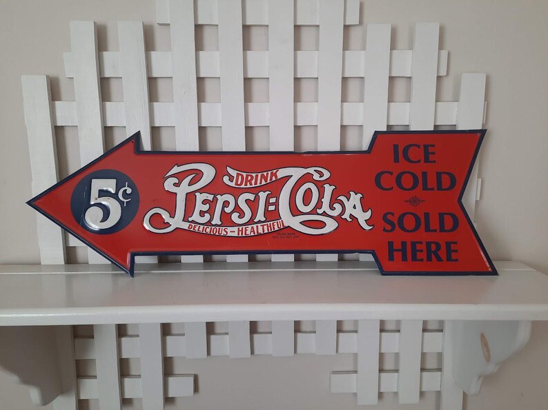 May include: Vintage Pepsi-Cola arrow-shaped sign in red with white and blue accents. The sign reads "Drink Pepsi-Cola, Delicious-Healthful" and "Ice Cold Sold Here." The sign also features the price "5&cent;".