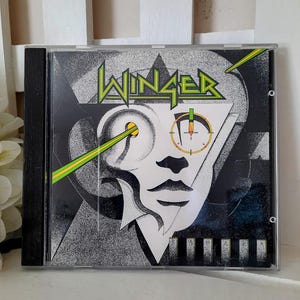 May include: A Winger music CD in a clear plastic jewel case. The cover art features a stylized black and white graphic of a face with a green laser beam pointing at the eye and a circular target sight. The word WINGER is printed in green.