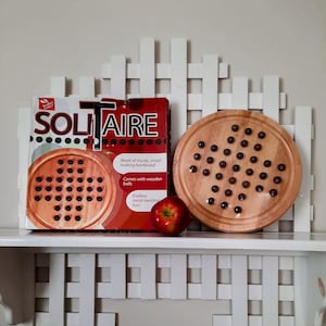 May include: A Solitaire board game set featuring a square cardboard box with red and white graphics, a round wooden game board with dark spherical pieces, and a single red apple placed between the box and the board.