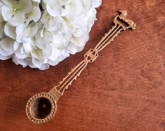 May include: A gold-toned metal spoon with a circular bowl featuring a beaded rim. The handle is long and slender with an openwork design, decorative notched edges, and a small horse-shaped finial at the very top of the handle.
