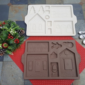 May include: Two gingerbread house molds, one white and one brown, with various shapes for creating gingerbread houses. A festive wreath with red berries and pinecones sits nearby, along with a set of measuring spoons.