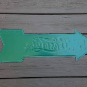 May include: A green metal arrow sign with embossed text. The sign is shaped like an arrow pointing to the right. The text on the sign reads "Taste Of". The sign is placed on a wooden surface.