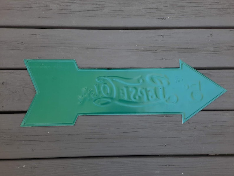 May include: A green metal arrow sign with embossed text. The sign is shaped like an arrow pointing to the right. The text on the sign reads "Taste Of". The sign is placed on a wooden surface.