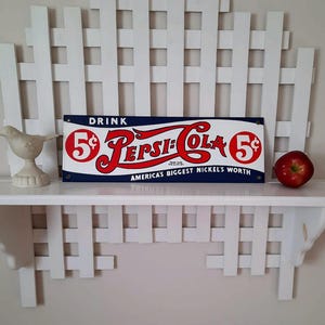 May include: A vintage Pepsi-Cola sign with red and white lettering on a blue background, advertising "America's Biggest Nickel's Worth." A white ceramic bird and a red apple sit on a white shelf, with a white lattice backdrop.