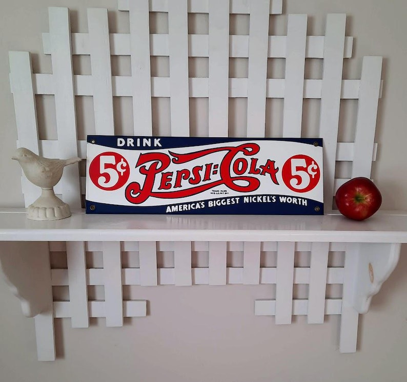 May include: A vintage Pepsi-Cola sign with red and white lettering on a blue background, advertising "America's Biggest Nickel's Worth." A white ceramic bird and a red apple sit on a white shelf, with a white lattice backdrop.