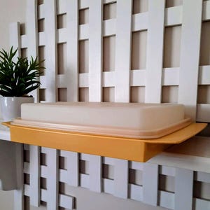 May include: A vintage yellow rectangular container with a cream-colored lid. The container is sitting on a white shelf in front of a white lattice background. A small potted plant is to the left of the container.