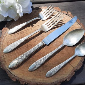 May include: Five pieces of silverware, including two forks, a knife, and two spoons, are arranged on a wooden surface. The silverware is silver-toned and has an ornate design.