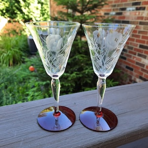 May include: Two clear glass wine glasses with etched floral designs and red bases. The glasses are on a wooden surface.