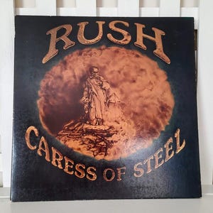 May include: A vinyl record album cover with the band name "RUSH" in gold lettering at the top. The album title "CARESS OF STEEL" is in gold at the bottom. The artwork features a figure in a robe standing on a rocky surface, set against a fiery orange and brown background.