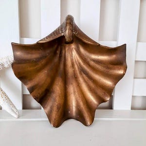 May include: A bronze-colored, seashell-shaped decorative dish. The dish has a textured surface and a detailed, raised design at the top. The dish is displayed on a white surface, suggesting a coastal or nautical theme.