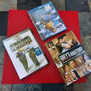 May include: Three DVDs and Blu-ray discs on a red woven placemat. Titles include "The Producers," "Monty Python and the Holy Grail," and "Grey's Anatomy: Complete Fifth Season More Moments."