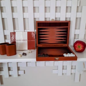 May include: A brown leather-like backgammon set with an open board featuring black and brown points. The set includes two brown dice cups, white and black checkers, dice, a doubling cube, and a manual with the text BACKGAMMON on the cover.