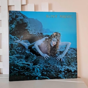 May include: Roxy Music album cover featuring a woman with curly hair and red lipstick, posed on a rocky surface. The album title is in gold lettering against a blue background. The overall aesthetic is artistic and evocative.