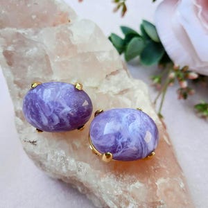 May include: A pair of oval-shaped stud earrings. Each earring features a marbled purple stone set in a gold-colored metal frame. The earrings are displayed on a light pink, textured surface, with floral accents in the background.