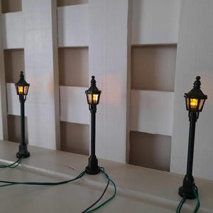 May include: Three miniature black street lamps with glowing yellow lights. Each lamp features a classic lantern-style head with clear panes and a thin black post. Thin green and black electrical wires extend from the base of each lamp.