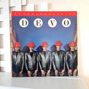 May include: Album cover for Devo's "Freedom of Choice." The cover features five figures in silver jumpsuits and red, geometric hats against a blue backdrop with the band's name and the album title.