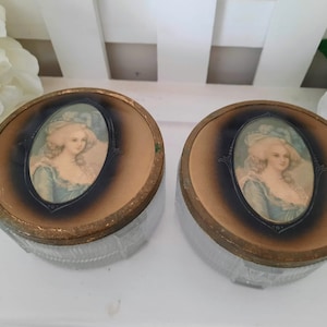 May include: Two vintage glass and gold-rimmed powder compacts with a portrait of a woman in a blue dress and white hat on the lid.
