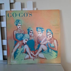May include: Album cover for "Beauty and the Beat" by Go-Go's. The cover features five women in teal towels and head wraps, with white face makeup and red lipstick. The album title is in teal, and the album name is in a peach and orange background.