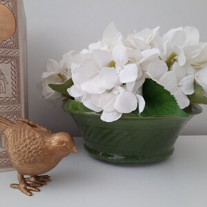 May include: A gold-colored bird figurine with a textured surface sits on a white surface in front of a green planter with white hydrangeas.