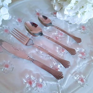 May include: A set of four silver-toned cutlery pieces, including a fork, knife, and two spoons, arranged on a white background with pink floral designs.