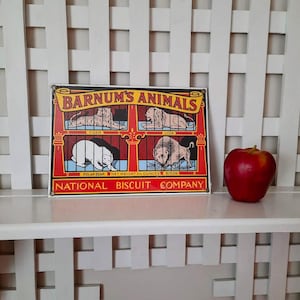 May include: A vintage metal sign with the text "Barnum's Animals" featuring illustrations of a tiger, lion, polar bear, and lion. A red apple sits to the right of the sign on a white shelf. The sign is from the National Biscuit Company.