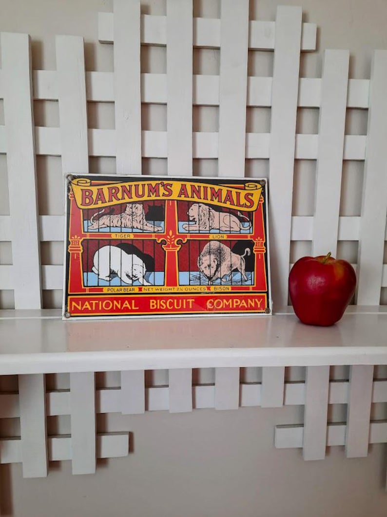 May include: A vintage metal sign with the text "Barnum's Animals" featuring illustrations of a tiger, lion, polar bear, and lion. A red apple sits to the right of the sign on a white shelf. The sign is from the National Biscuit Company.