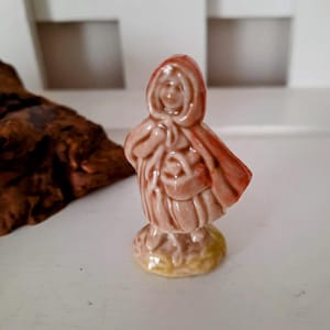 May include: A small ceramic figurine of a girl wearing a red hooded cloak and holding a basket. The figurine is painted in shades of brown, red, and yellow.