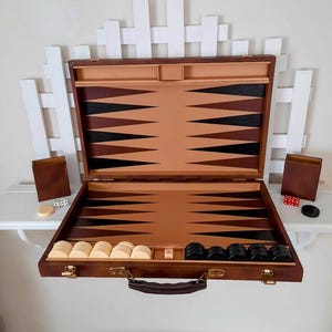 May include: Open brown backgammon set with light brown and black triangles. The set includes white and black game pieces, dice, and two brown leather dice cups. The set is displayed on a white shelf.
