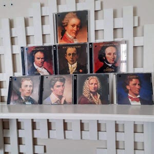 May include: A collection of compact discs featuring portraits of classical composers. The CDs are arranged on a white shelf and include composers such as Mozart, Beethoven, and Chopin. Each CD case displays the composer's name.