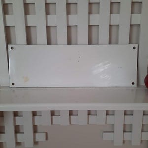 May include: A rectangular white metal sign with rounded corners, mounted on a white shelf. The sign has four visible screw holes and some minor discoloration. The shelf is part of a white, lattice-style structure.