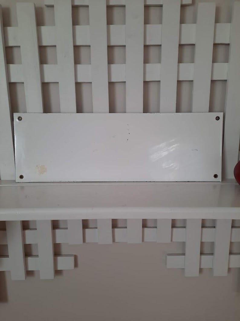 May include: A rectangular white metal sign with rounded corners, mounted on a white shelf. The sign has four visible screw holes and some minor discoloration. The shelf is part of a white, lattice-style structure.