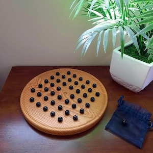 May include: A round wooden board game featuring a grid of small indentations filled with smooth black marbles. A small navy blue drawstring pouch sits next to the board, containing one additional black marble, completing the set for play.