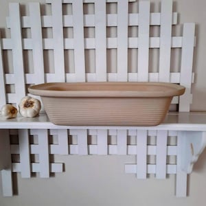 May include: A beige, rectangular ceramic baking dish with a ribbed texture, resting on a white shelf. Two garlic bulbs are placed to the left of the dish. The shelf is part of a white, lattice-style wall decor.