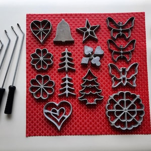 May include: A collection of gray metal cookie cutters in various shapes, including hearts, stars, butterflies, and Christmas trees, arranged on a red textured surface. Four metal tools with black handles are also present.
