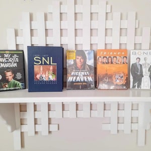 May include: A white shelf displays five DVD cases. Titles include "My Favorite Martian," "SNL," "Highway to Heaven," "Friends," and "Bones." The shelf is part of a white, decorative wall feature with a lattice design.