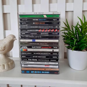May include: A stack of CDs featuring various artists and albums, including ZZ Top, The Who, U2, Santana, Fleetwood Mac, Eurythmics, and The Cars. The CDs are in clear plastic cases, with album titles and artist names visible. A decorative bird and a potted plant are in the background.